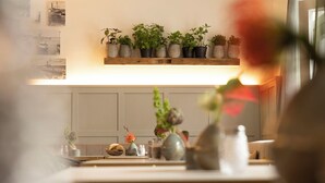 A kitchen with a wooden shelf holding potted plants, a dining table with a vase of flowers, and a wall with framed photographs. - Boutiquehotel Petuh am Hafen - adults only (Flensburg)