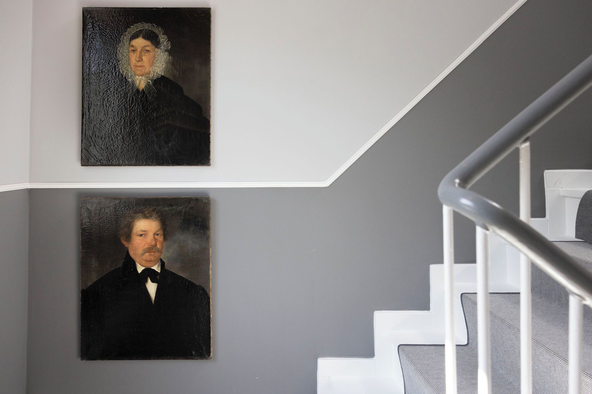Two portraits on a wall, a staircase with white railings, and a grey wall.