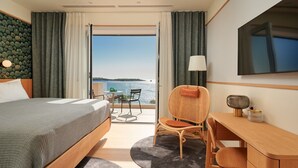 A hotel room with a bed, a desk, a chair, a TV, and a view of the sea.