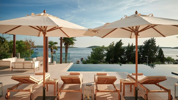 A poolside area with lounge chairs and umbrellas, overlooking a body of water and greenery.