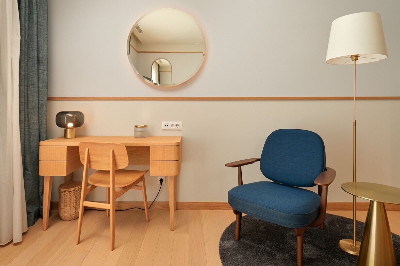 Une pièce comprenant un bureau en bois, une chaise bleue, un miroir rond, une lampe et une petite table ronde.