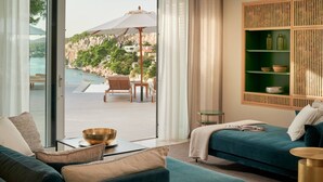 Une chambre d’hôtel moderne avec vue sur une piscine et des montagnes, un canapé turquoise, une étagère verte agrémentée d’objets décoratifs et une porte en verre donnant sur l’extérieur.