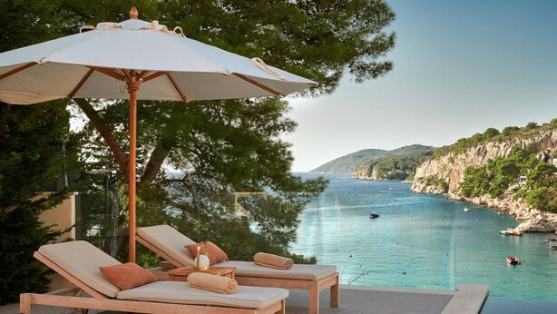 Un espace aménagé au bord de la piscine, avec deux chaises longues et un grand parasol, offrant une vue sur une baie côtière avec des bateaux et des falaises.