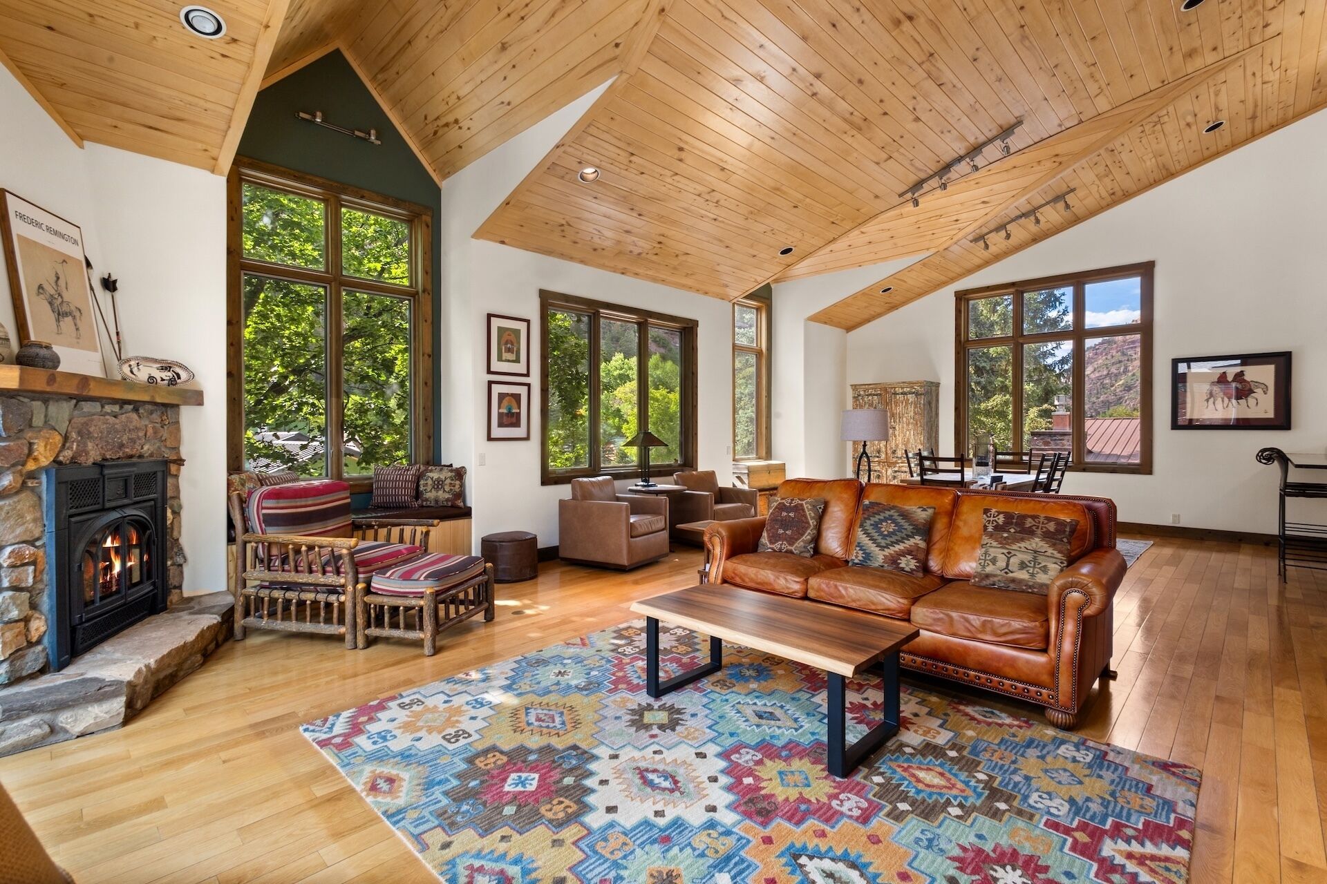 A spacious living room with a fireplace, leather sofa, wooden flooring, and large windows offering a view of the outdoors.