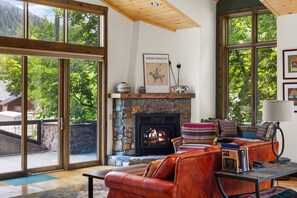 Una sala de estar acogedora con chimenea de piedra, sofá de cuero y grandes ventanas que ofrecen vistas a árboles y montañas.