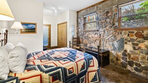 A bedroom with a stone fireplace, a quilted bedspread, and a view through a window. - Spacious Getaway | Walk to Downtown | Private Courtyard (Ouray)