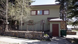 Un edificio de dos plantas con un muro de piedra y una reja metálica, un pequeño garaje adosado y una puerta roja.