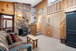 A cozy living room with a stone fireplace, wooden walls, a staircase, and a large cabinet.