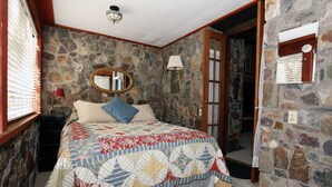 Un dormitorio con pared de piedra, cama con colcha estampada, espejo y mesita de noche con lámpara.