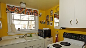 Una cocina con estufa blanca, un lavabo y una ventana con un adornado colorido. Hay armarios y un microondas en la encimera.
