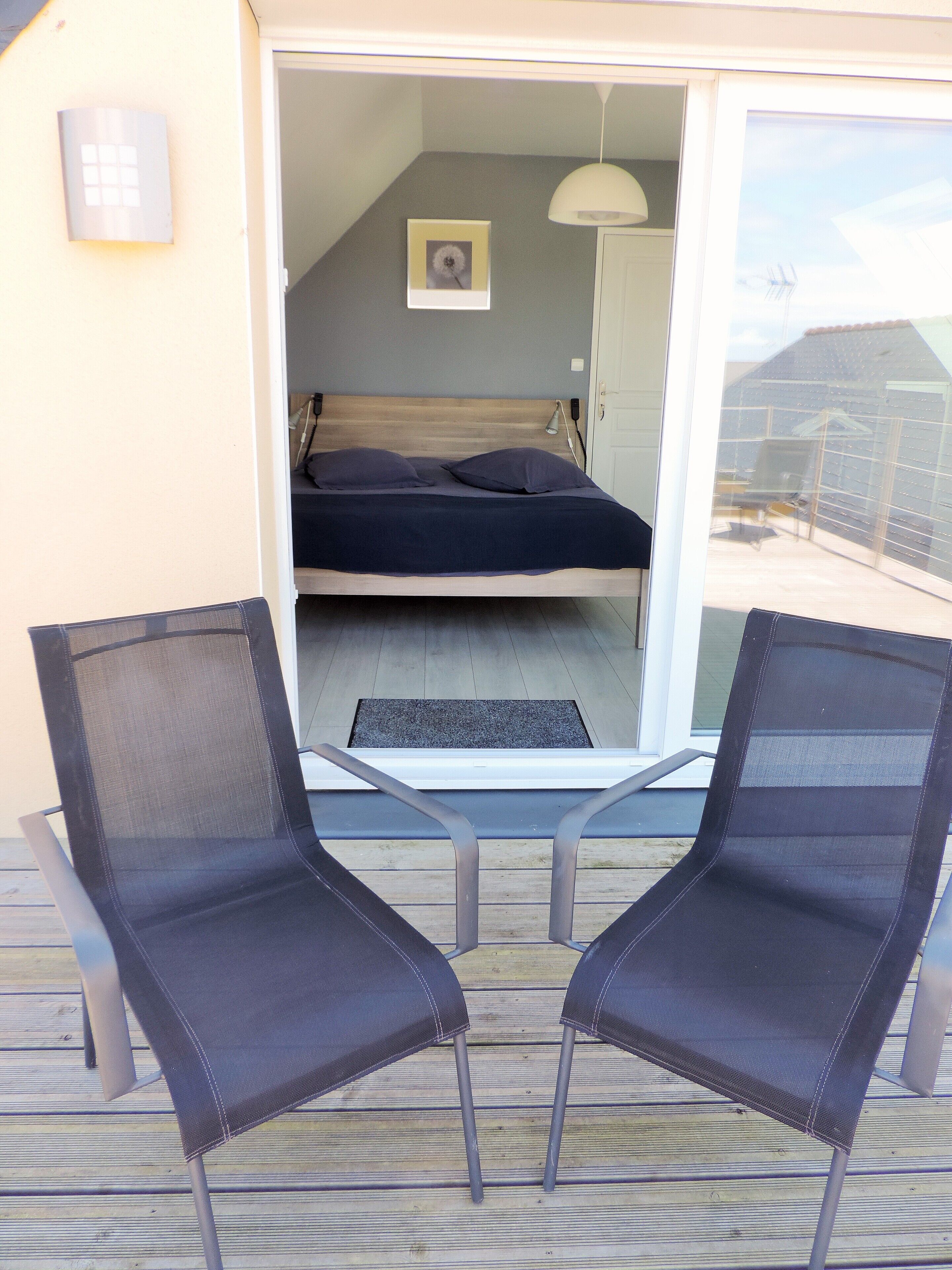 Ein Balkon mit zwei Stühlen, ein Bett mit dunkler Bettdecke, ein Bild an der Wand und eine weiße Tür.