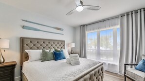 Een slaapkamer met een gebobbelde hoofdbord, een bed met witte beddengoed, een nachtkastje met een lamp, een stoel met een kussen, een plafondventilator en een raam met gordijnen.