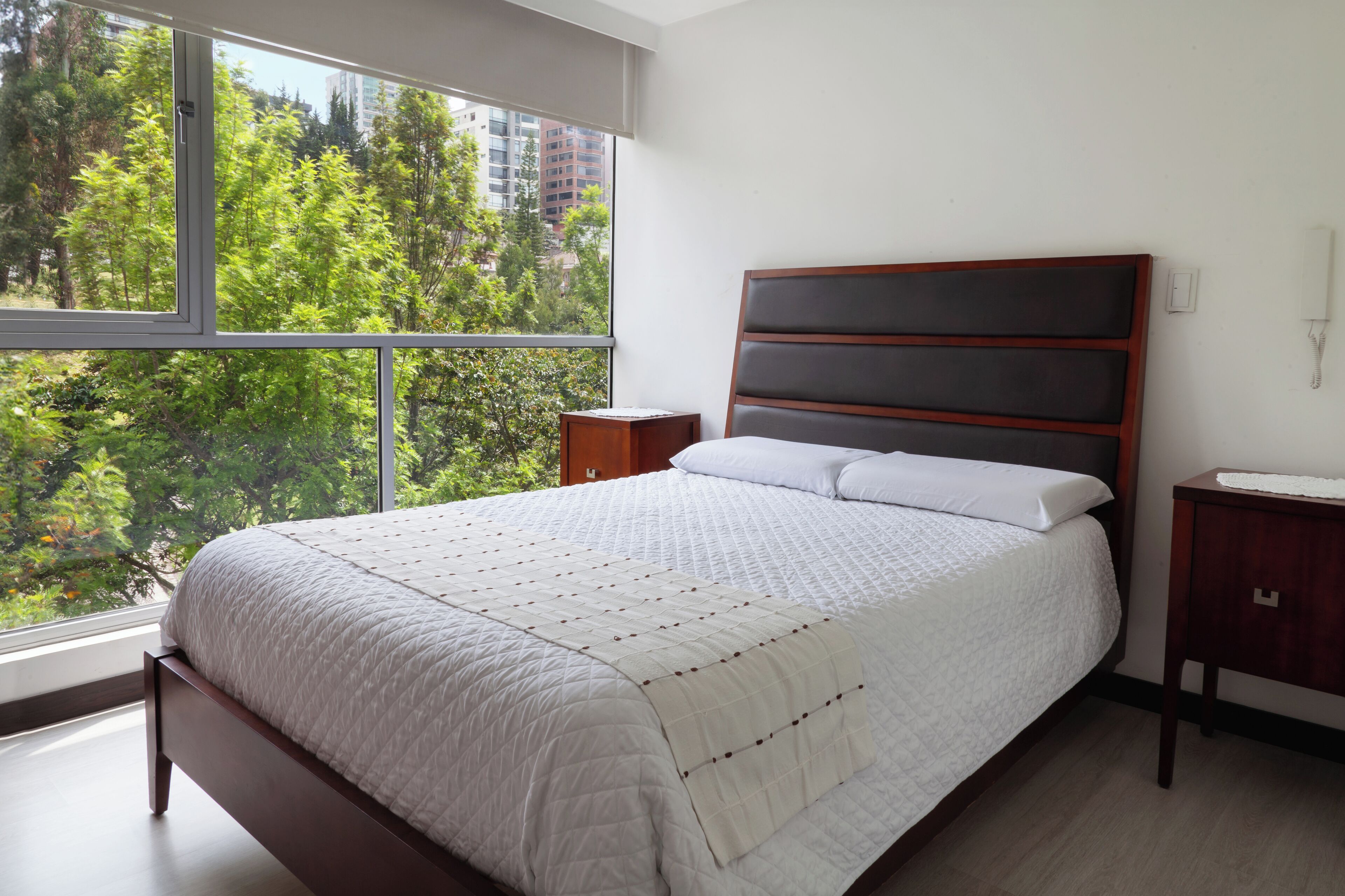Un dormitorio con un ventanal grande, una cama con cubrecama blanco acolchado, un cabecero de madera y una mesita de noche con lámpara.