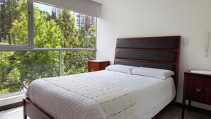 A bedroom with a large window, a bed with a quilted white bedspread, a wooden headboard, and a nightstand with a lamp. - 3BR La Carolina Quito Parking Sleeps 8 Galapagos Base 700Mbps (Quito)