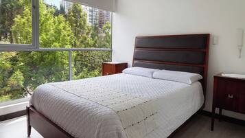 Un dormitorio con un ventanal grande, una cama con cubrecama blanco acolchado, un cabecero de madera y una mesita de noche con lámpara.