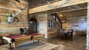 Un interior de madera con una mesa de billar, una chimenea y una escalera que conduce a un nivel superior.