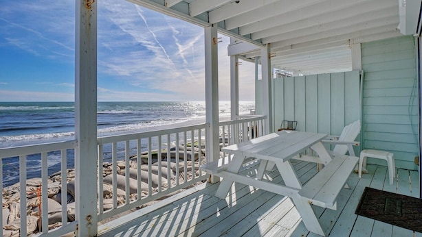 Una terraza de madera frente a la playa, con una mesa y sillas, con vista al océano.