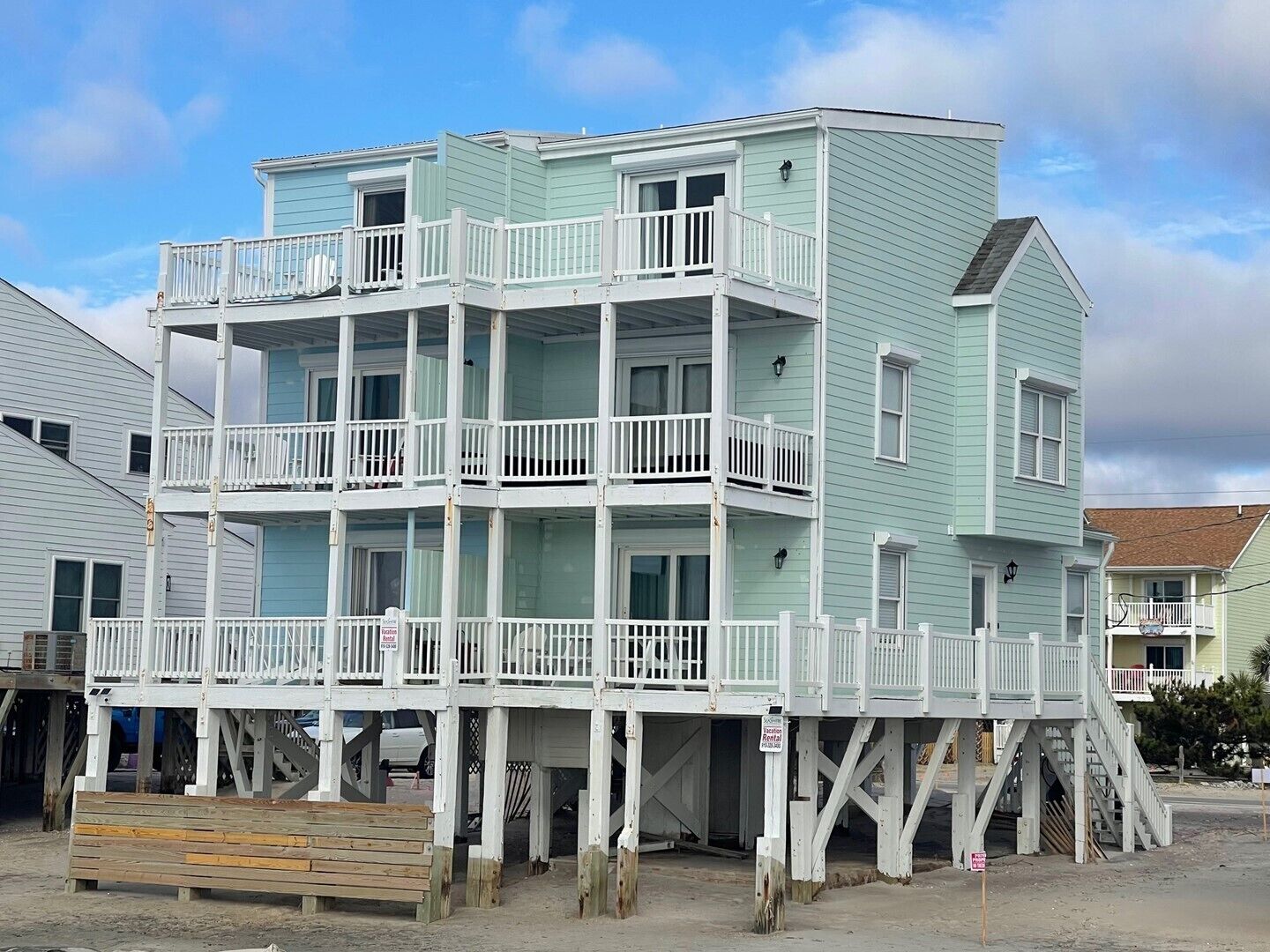 A multi-story beachfront building with balconies, a wooden deck, and a sign indicating it is a rental property.