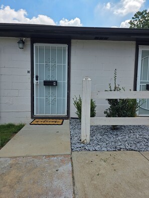 Ein Wohnhaus mit einem Metallgitter, einer Fußmatte mit dem Schriftzug 'welcome' und einem kleinen Gartenbereich mit Kies und Pflanzen.