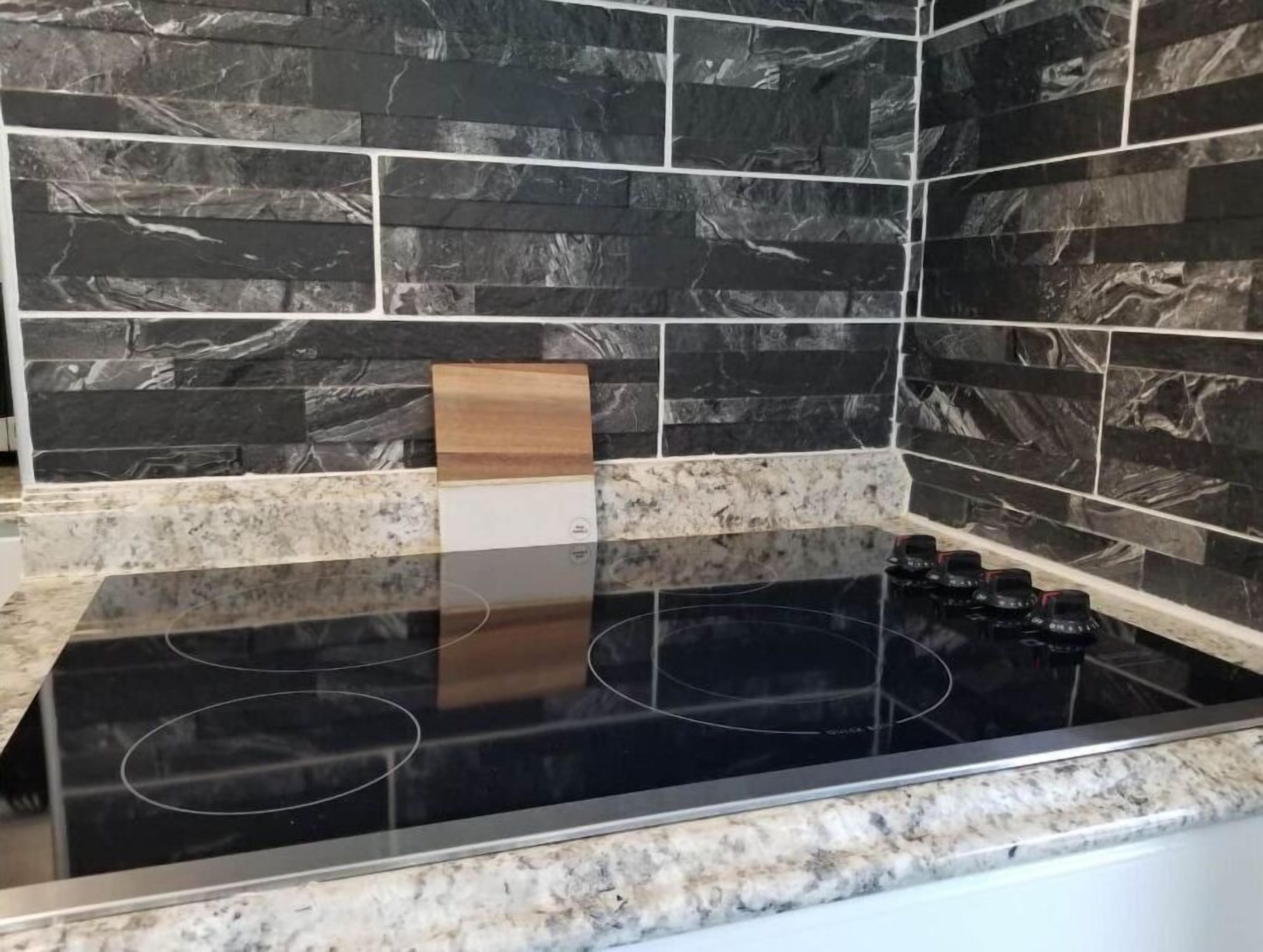A kitchen with a black stovetop, granite countertops, and a backsplash with dark, textured tiles.