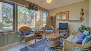 Una sala de estar acogedora con muebles de mimbre, televisor y ventanas amplias con vistas a árboles.