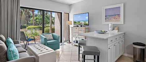 A modern living room with a sofa, ottoman, and a teal armchair. A kitchen area with a white island and bar stools is visible. A television is mounted on the wall, and a beach scene painting is displayed.
