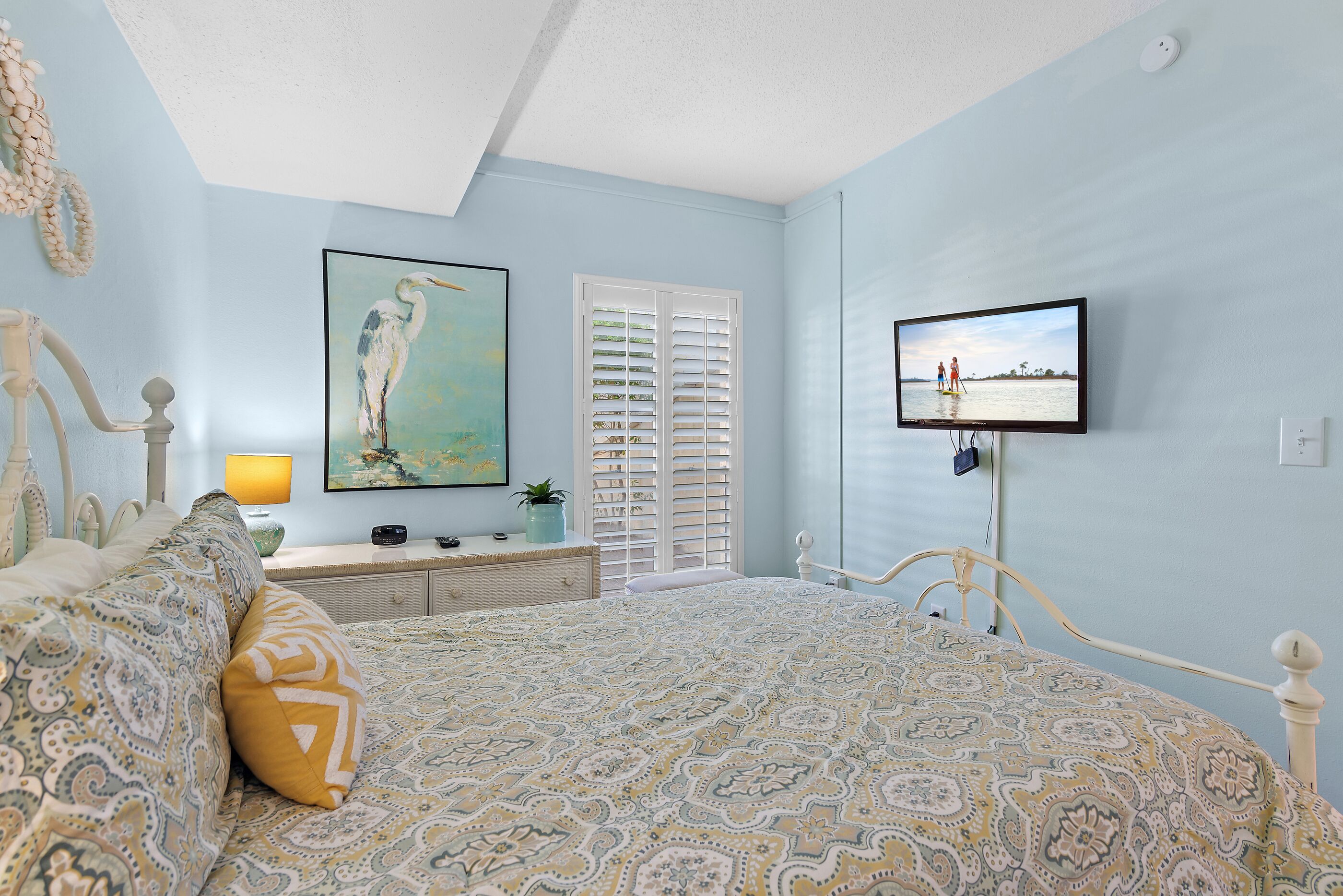 A bedroom with a bed, a television, a window with shutters, and a painting on the wall.