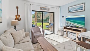 A living room with a sofa, armchair, wooden coffee table, TV, and a guitar. There's a sign reading "BEACH HOUSE" and a view of the outdoors.