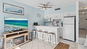 A modern kitchen with white cabinets, a built-in microwave, and a refrigerator. There is a bar area with stools and a television displaying a scenic view.