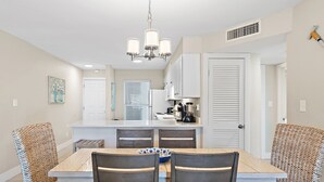 A dining area with a wooden table and wicker chairs, a chandelier, and a kitchen in the background. - Newly Renovated, unit 20 right on Beach… Just steps to the Sand (Destin)