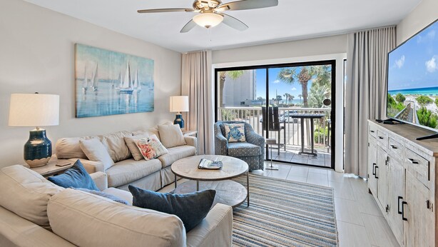 A modern living room with a sofa, coffee table, and a TV. There's a painting on the wall and a view of the outdoors through the glass door. - Newly Renovated, unit 20 right on Beach… Just steps to the Sand (Destin)