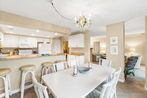 Une cuisine spacieuse avec des meubles blancs, un îlot central doté de tabourets, une table à manger avec des chaises et un lustre.