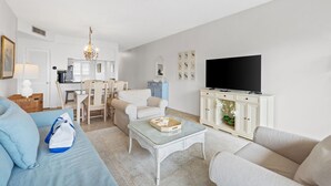 A living room with a sofa, a coffee table, a TV, and a dining area with chairs and a table. - Beach-side, beautiful low-rise with uncrowded beach! (Destin)