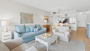 A living room with a blue sofa, white armchairs, and a coffee table. A kitchen is visible in the background. - Beach-side, beautiful low-rise with uncrowded beach! (Destin)