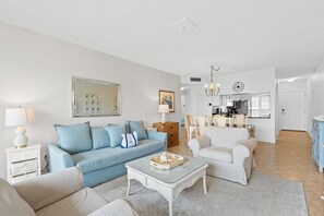 Sala de estar com um sofá azul, poltronas brancas e uma mesa de centro. Cozinha visível ao fundo.