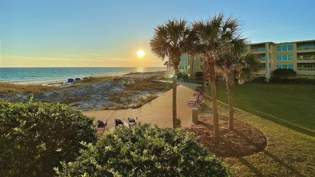 A beachfront hotel with a paved walkway, palm trees, and a view of the ocean at sunset. - Beach-side, beautiful low-rise with uncrowded beach! (Destin)