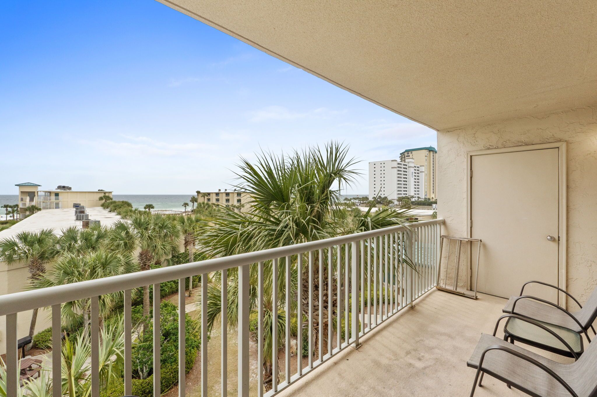 A balcony with a view of the beach and palm trees, a door, and a chair.