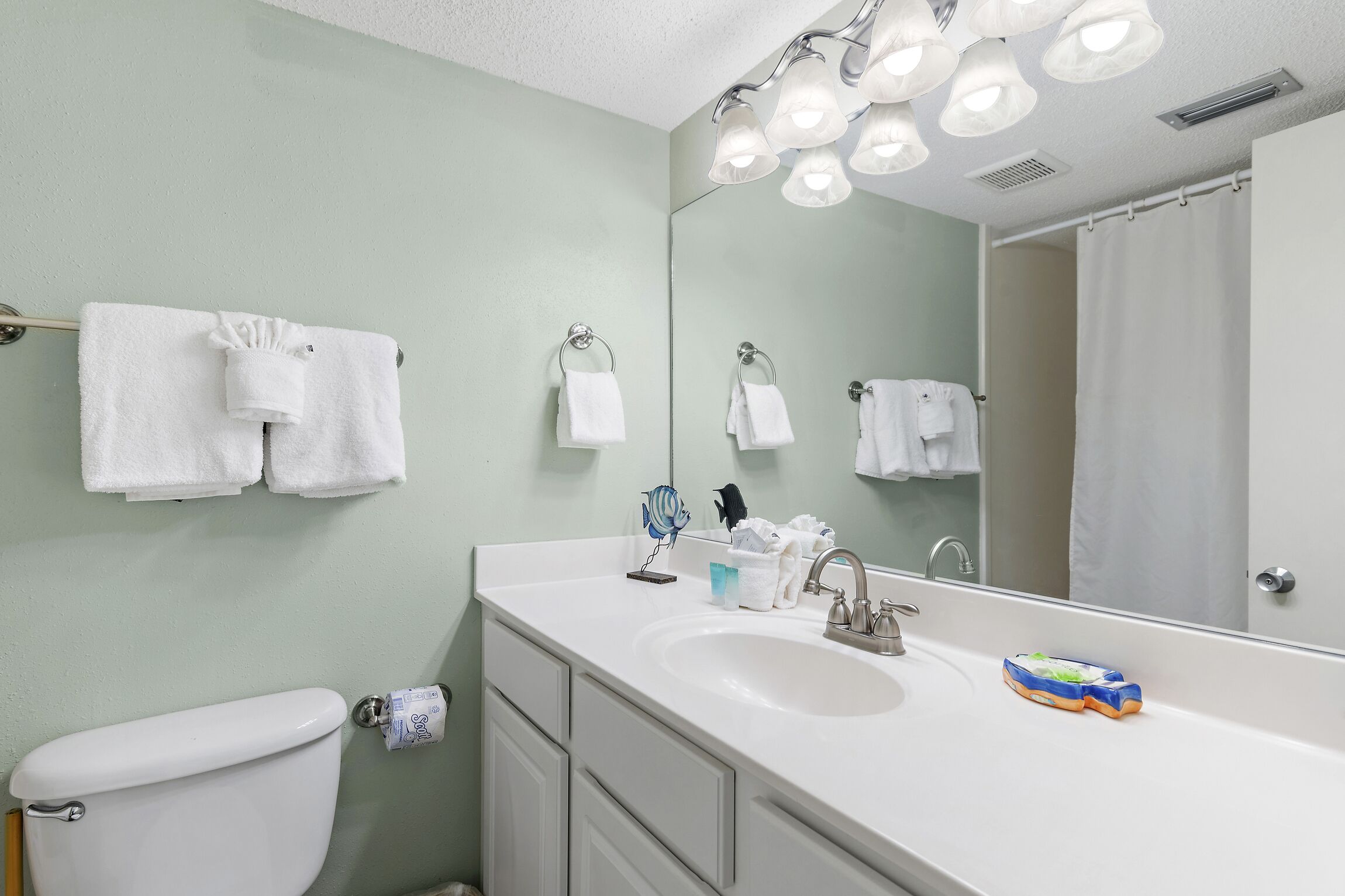 Une salle de bain avec un lavabo blanc, un miroir et des toilettes. On peut voir des serviettes accrochées au mur et un rideau de douche qui se reflète dans le miroir.