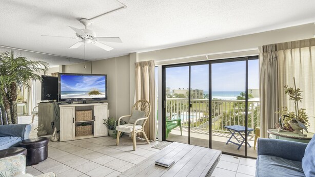 Een ruime woonkamer met een grote televisie, comfortabele zitplaatsen, een balkon met uitzicht op het strand en een plafondventilator.