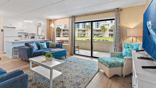 Un salon moderne avec un canapé, un fauteuil et une table à manger. On aperçoit une cuisine au fond, et un balcon avec vue sur les bâtiments est également visible.