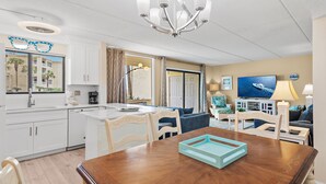 Une pièce bien éclairée, comprenant une table à manger, des chaises, un lustre, un téléviseur et un coin cuisine.