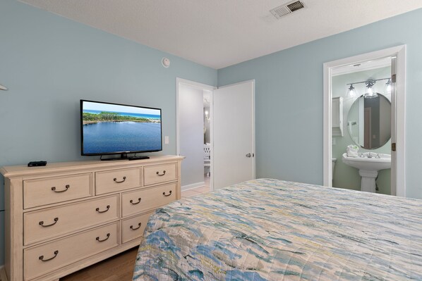 A bedroom with a bed, a dresser, a television displaying a scenic image, a bathroom with a sink and mirror, and a door leading to another room. - Beach-side, beautiful low-rise with uncrowded beach! (Destin)
