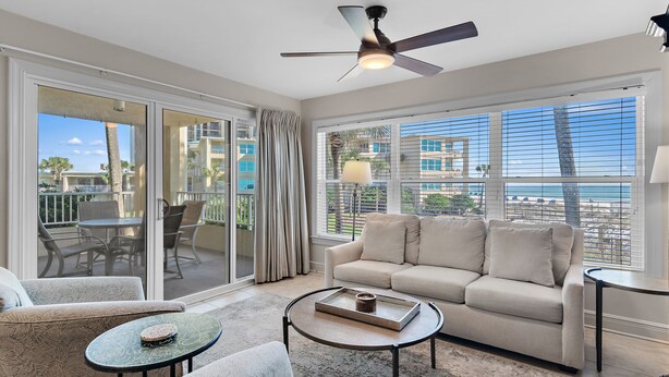 A spacious living room with a beige sofa, a coffee table, and a ceiling fan. Large windows offer a view of the beach and buildings. - Beach-side, beautiful low-rise with uncrowded beach! (Destin)