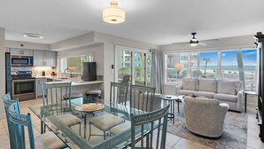 A modern kitchen with stainless steel appliances, a dining area with a glass table and blue chairs, and a living room with a sofa and a view of the ocean. - Beach-side, beautiful low-rise with uncrowded beach! (Destin)