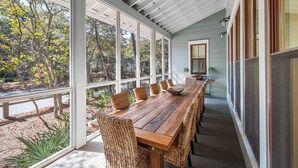 A covered patio with a long wooden table and wicker chairs, large windows, and a view of trees.