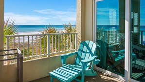 Ein Balkon mit Blick auf den Strand, ein türkisner Adirondack-Stuhl und eine Glastür zu einem Balkon mit Geländer.