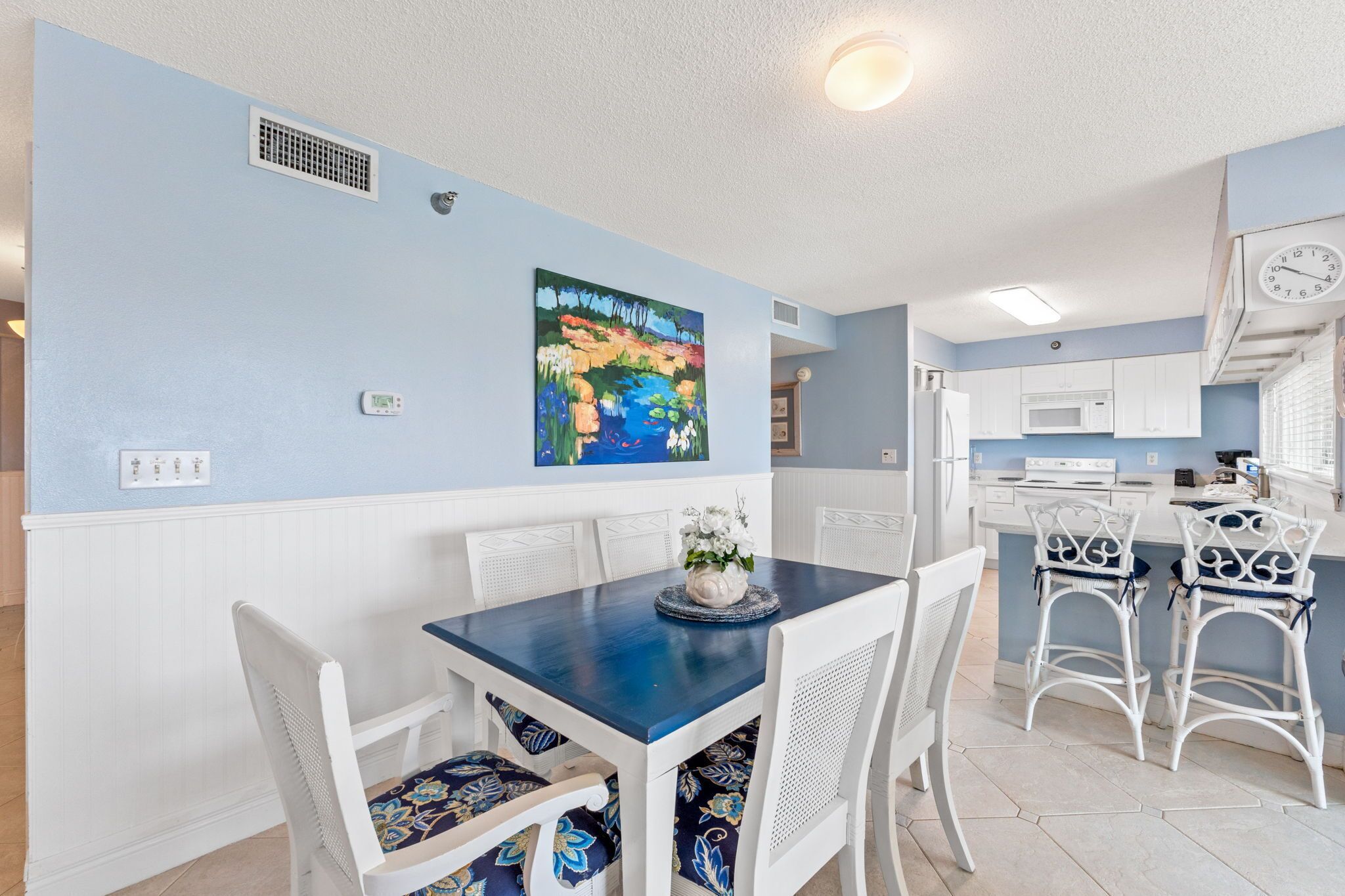 A dining area with a blue table and white chairs, a painting on the wall, and a kitchen in the background.