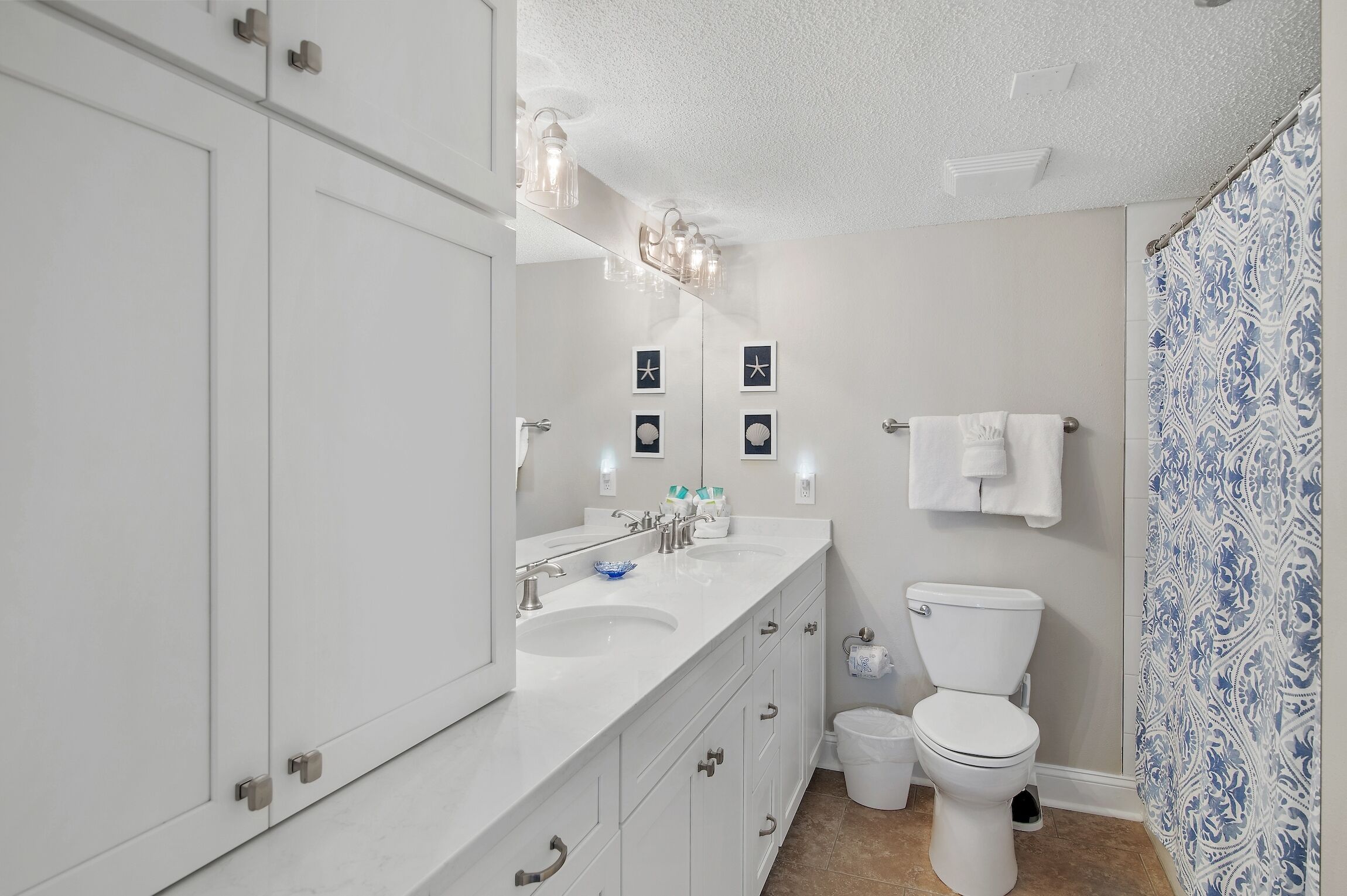 A modern bathroom with white cabinetry, a double sink, a toilet, and a shower with patterned curtains.