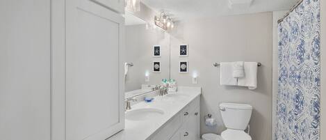 A modern bathroom with white cabinetry, a double sink, a toilet, and a shower with patterned curtains.