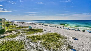 A beach with a row of umbrellas and beach chairs, a building with a balcony, and a clear blue sky. - TOP FLOOR GULF FRONT UNIT!! B1404 PANAROMIC VIEW OF GULF OF MEXICO! (Destin)